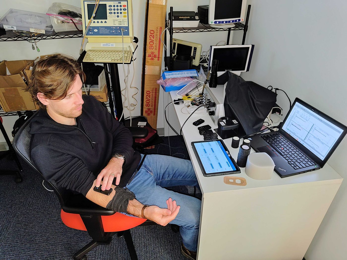 Thibault at a lab desk wearing a sensor band on his upper arm, with live signals on a laptop and a tablet
