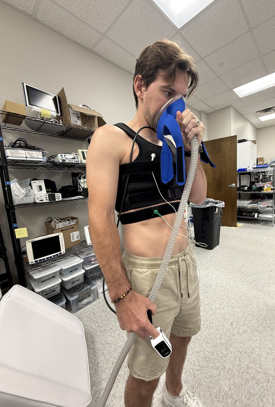 Thibault breathing through a mask and hose while wearing a chest sensor harness and holding a finger pulse oximeter. Breathing low-oxygen air through a mask, with chest sensors and a reference finger pulse oximeter.