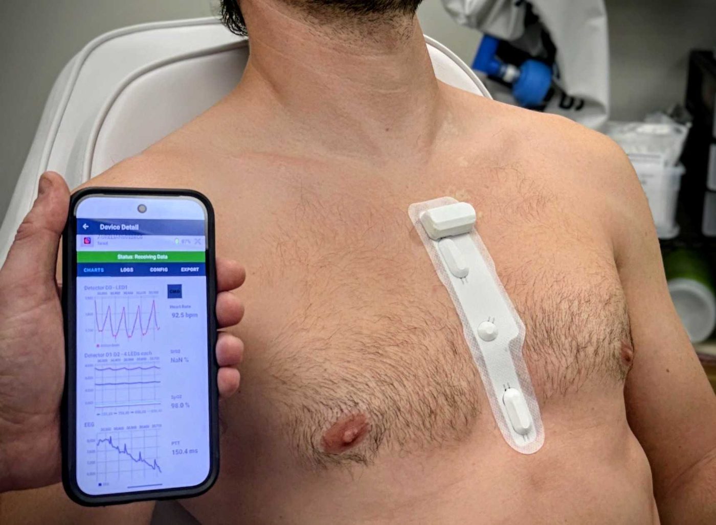 A thin sensor patch on the upper chest next to a phone held up to the camera. A chest sensor patch streaming to a phone app.