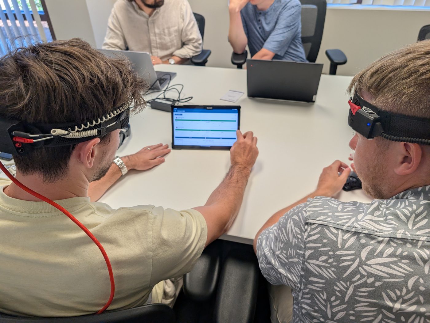 Two people wearing sensor headbands at a conference table, one tapping a tablet. Trying brain-sensing headbands with the team.